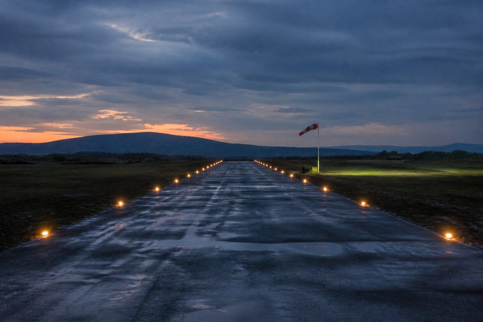 A single narrow asphalt airstrip at dusk receding towards low hills, with edge lights just switched on in two converging lines and a windsock on a pole in the middle distance.