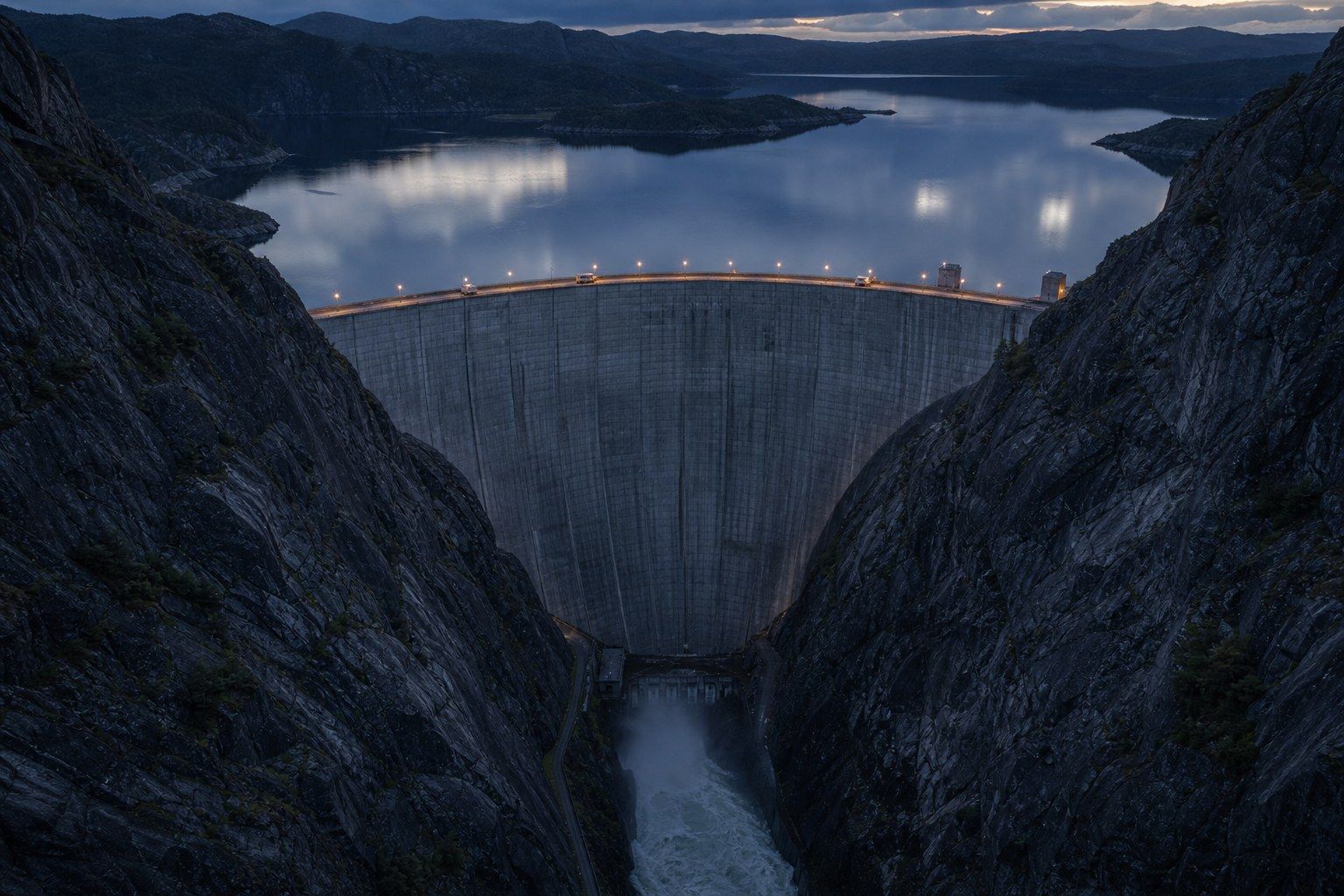 Aerial view at dusk of a large concrete arch dam curving between two steep granite gorge walls, with a lit service road along the crest and a pale turbulent tailrace channel far below.