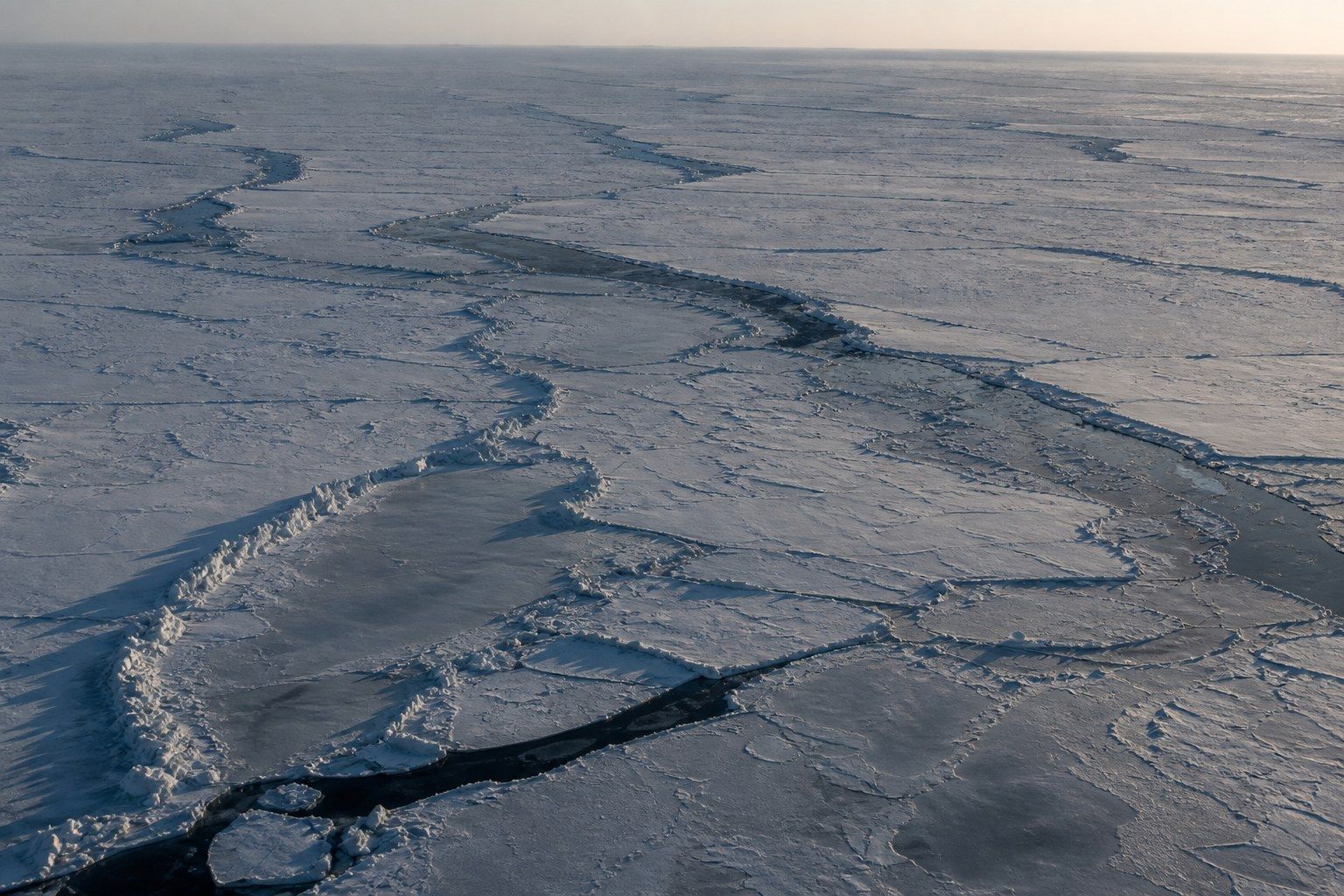 Aerial view of Arctic pack ice in late winter under a very low sun, broken by long dark open-water leads with pressure ridges casting long blue shadows.