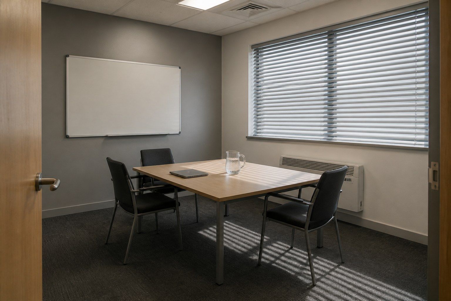 A small empty meeting room set up for an interview seen from the doorway, with a plain table, two chairs on one side and one on the other, a closed laptop and venetian blinds half closed.