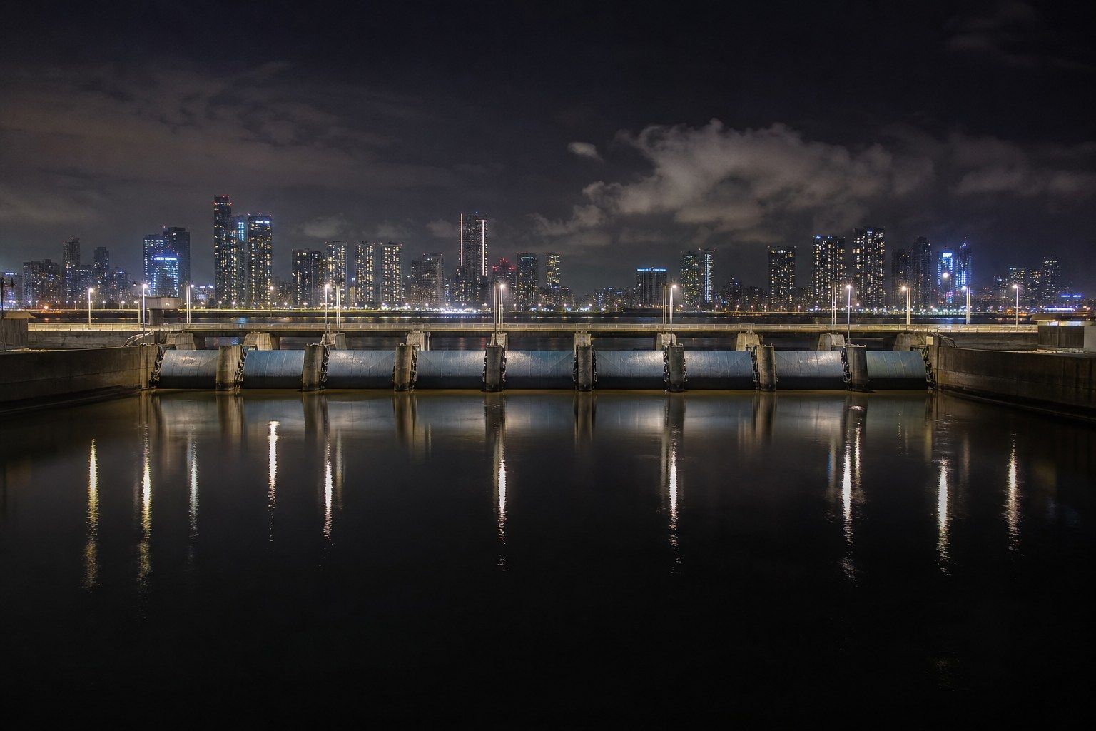 A row of steel crest gates spanning a wide channel at night, with a lit city skyline on the horizon behind and long reflections of lamp posts on still black water.