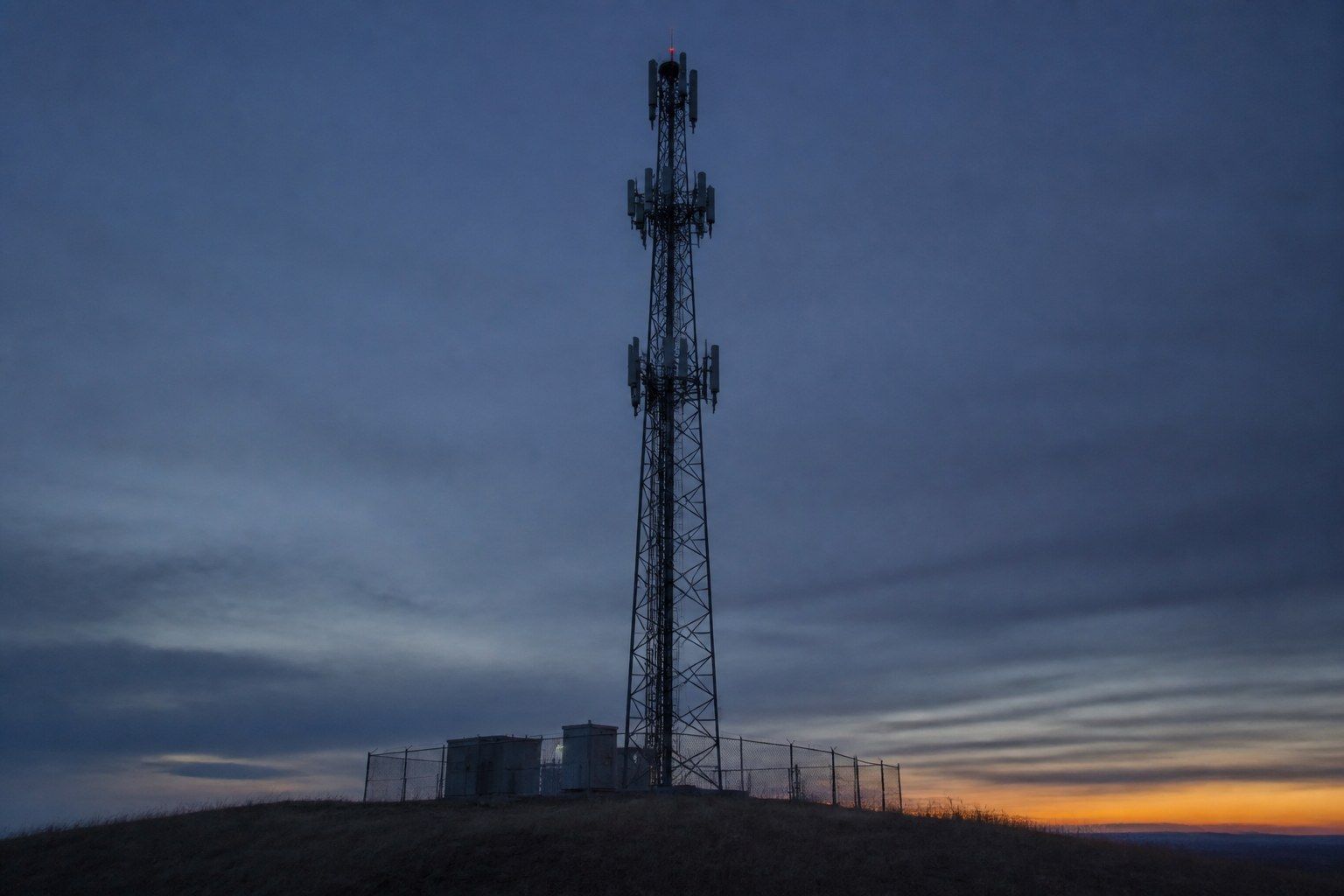 A lattice telecommunications mast on a low hill at dusk seen from below against a deep blue-grey sky, with panel antennas at three levels and small equipment cabinets in a chain-link compound at its base.