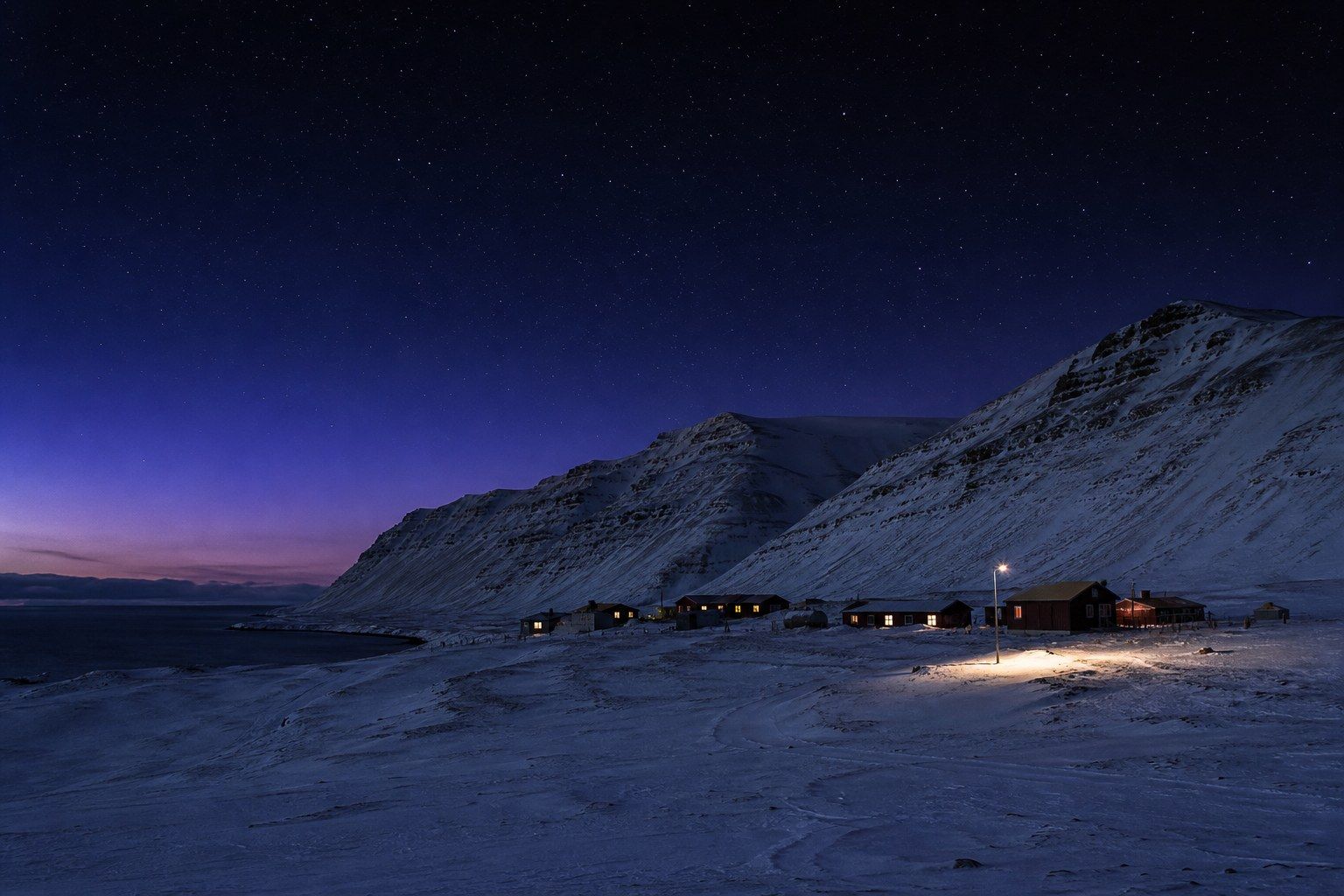 A small Arctic coastal settlement of dark low buildings with lit windows beneath steep snow-covered mountains, photographed at midday under a narrow band of deep indigo twilight low on the horizon.
