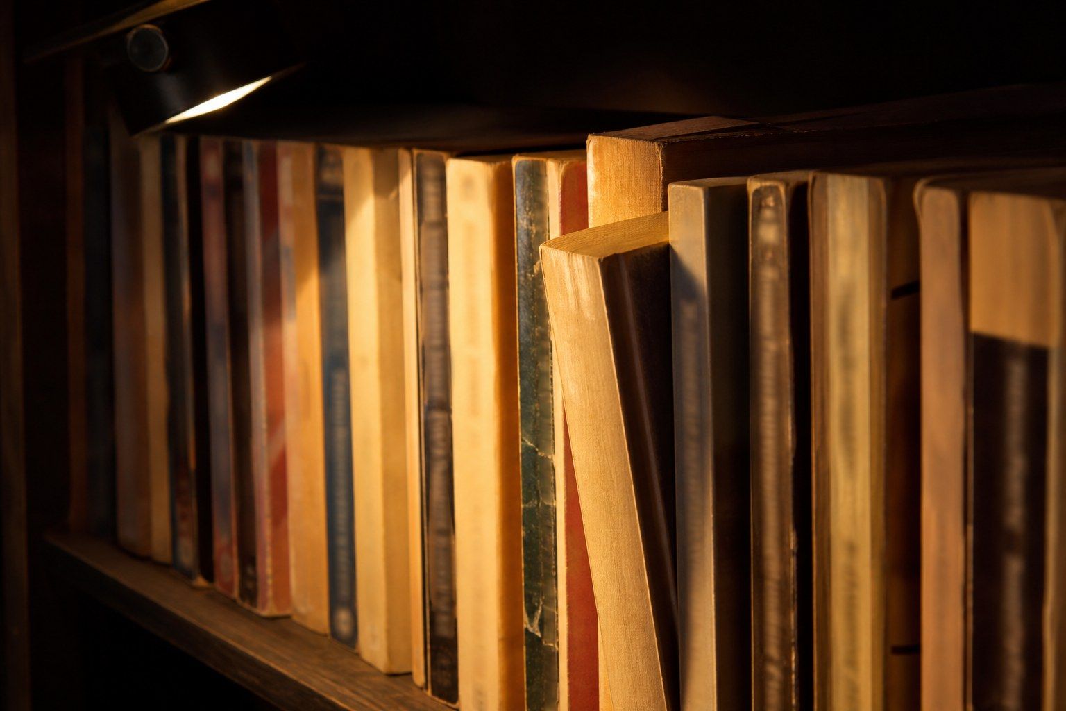 A tightly packed shelf of well-read mass-market paperback novels under a low warm reading lamp in a dark room, spines cracked and creased with edges yellowed.