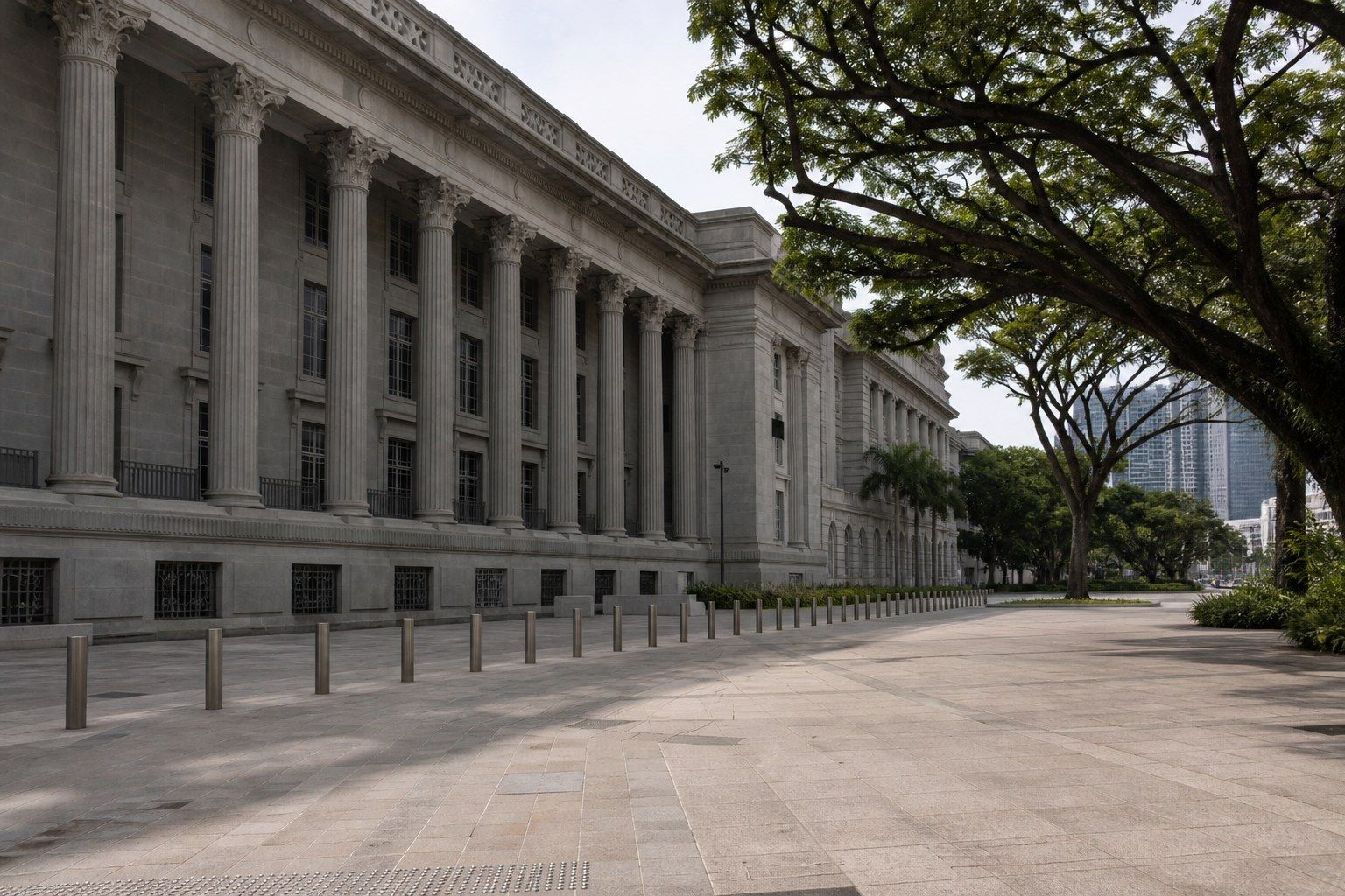 A neoclassical stone government building with a colonnade in the Singapore civic district, mature rain trees and an empty forecourt in late morning light.