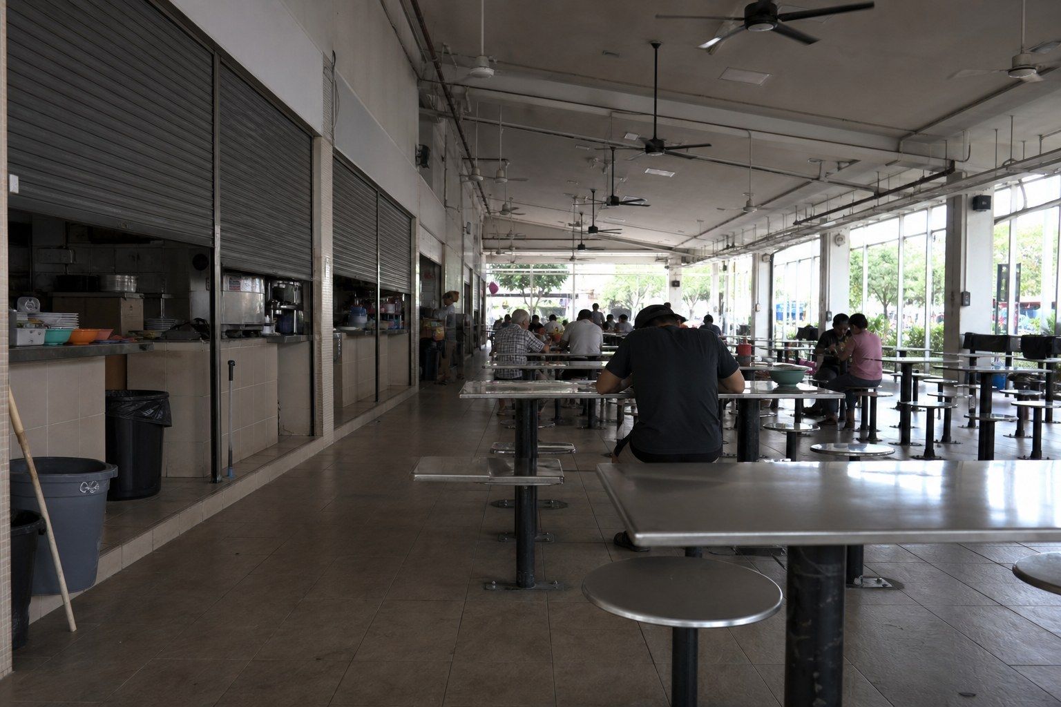 The interior of a Singapore hawker centre in mid-afternoon, rows of steel tables and stools bolted to the floor under ceiling fans, stall shutters half down.