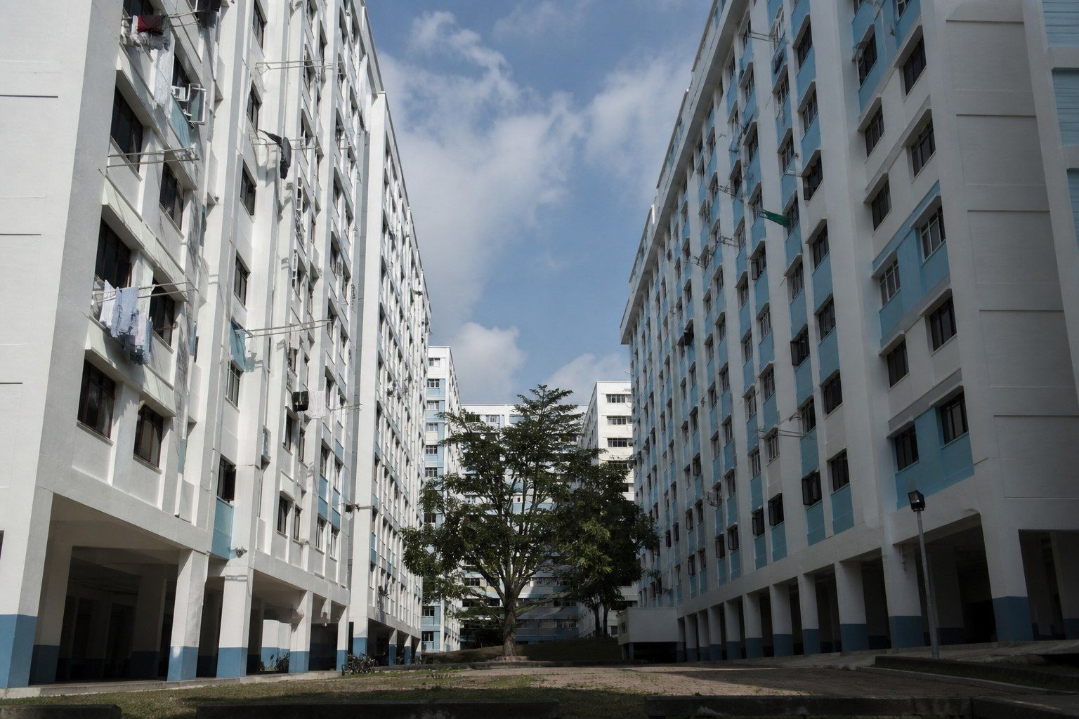 Two long slab blocks of Singapore public housing seen from ground level, laundry poles and window awnings above void deck pillars in hard afternoon light.