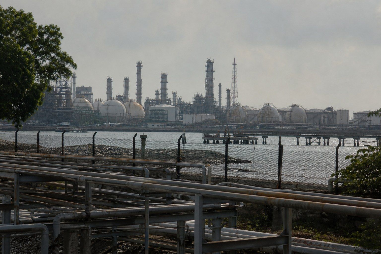 An industrial waterfront in western Singapore, refinery towers and spherical storage tanks behind a fence with pipework racks in the foreground.