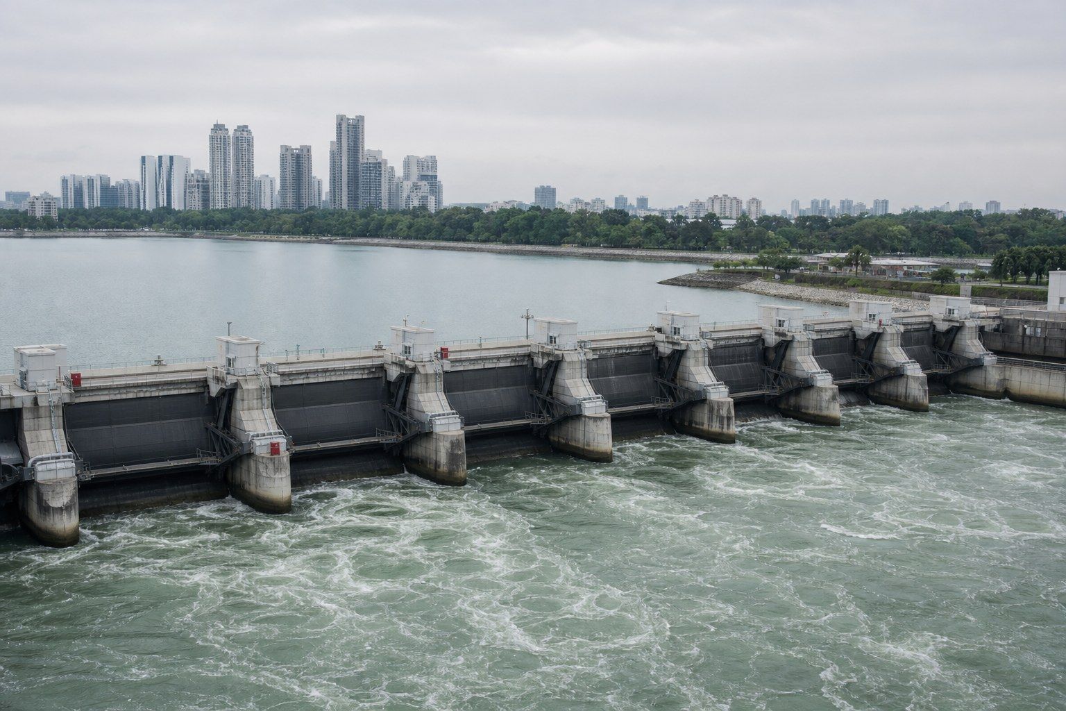 A tidal barrage across a river mouth in Singapore, steel crest gates set in concrete, a freshwater basin on one side and the sea on the other, city towers behind.