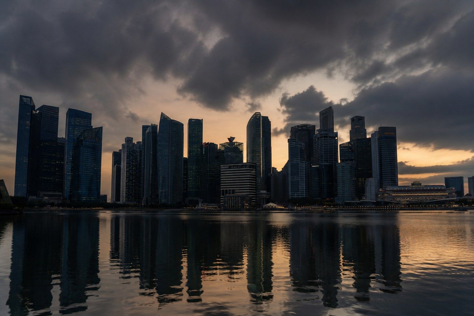 The Singapore central business district across Marina Bay at dusk, office towers in silhouette under monsoon cloud with their windows reflected in still water.