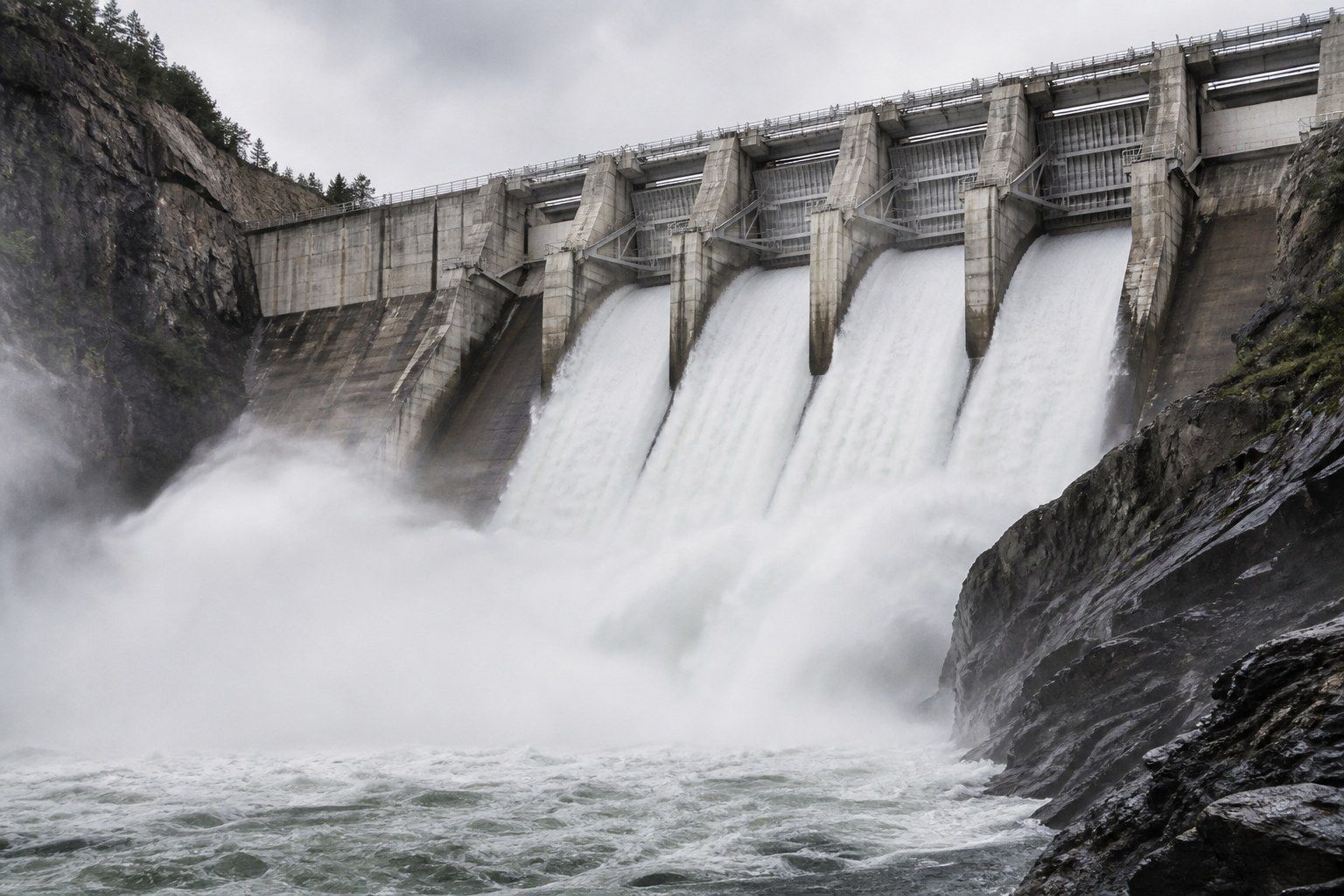 A large concrete gravity dam seen from downstream at midday with several spillway bays open, discharging white turbulent water in broad plumes with spray drifting across dark wet canyon rock.