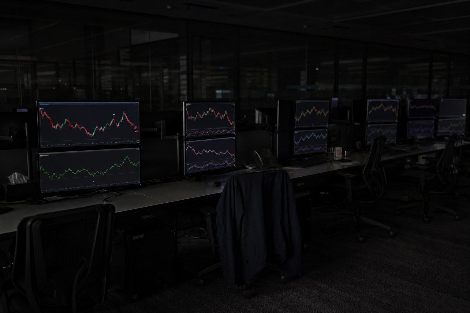 An empty institutional trading desk at night with a long row of dark workstations, several monitors still displaying abstract candlestick and line charts in muted red and green with no readable numbers.